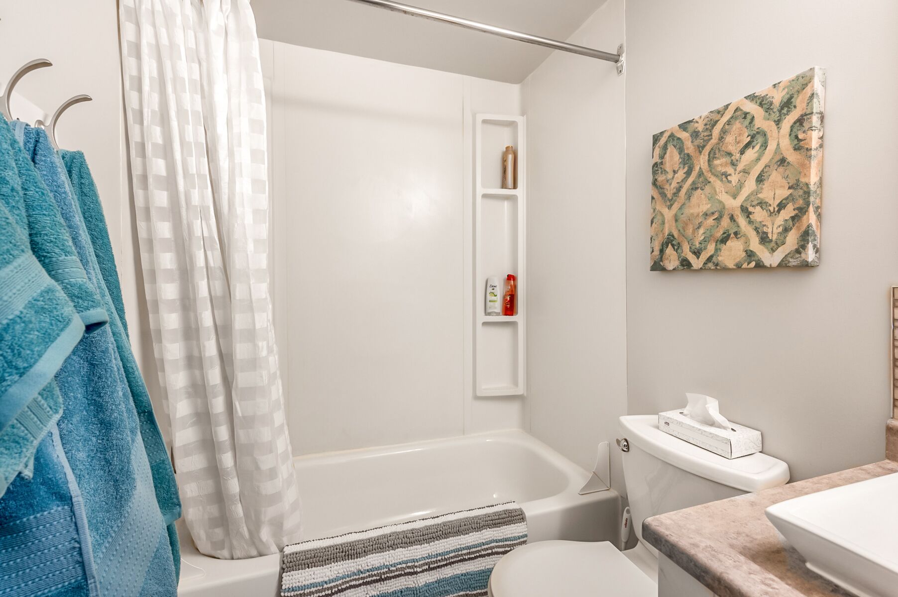 A bathroom with a bathtub, a toilet, a towel rack with blue towels, a shower curtain, a shelf with toiletries, and a patterned wall art piece.