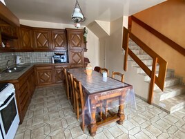 Una cocina con armarios de madera, salpicadero de azulejos, una mesa de comedor con sillas, una lámpara colgante y una escalera.