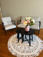 Una habitación con un tapete redondo con flecos, dos sillas, un sofá y una mesa pequeña con un florero.