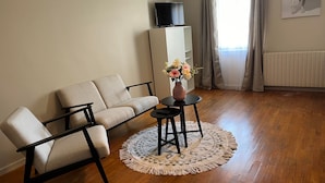 A living room with a sofa, a coffee table with a vase of flowers, a TV on a cabinet, and a radiator. - Gite le Saint André (Villers-sur-Port)