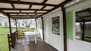 Eine überdachte Terrasse mit Holzdeck, einem weißen Tisch und Stühlen und Blick auf eine Wohnwagenanlage im Hintergrund.