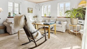 Un salon chaleureux avec un fauteuil à bascule, un canapé et une table basse. On y trouve des fenêtres, un lustre et une plante en pot.