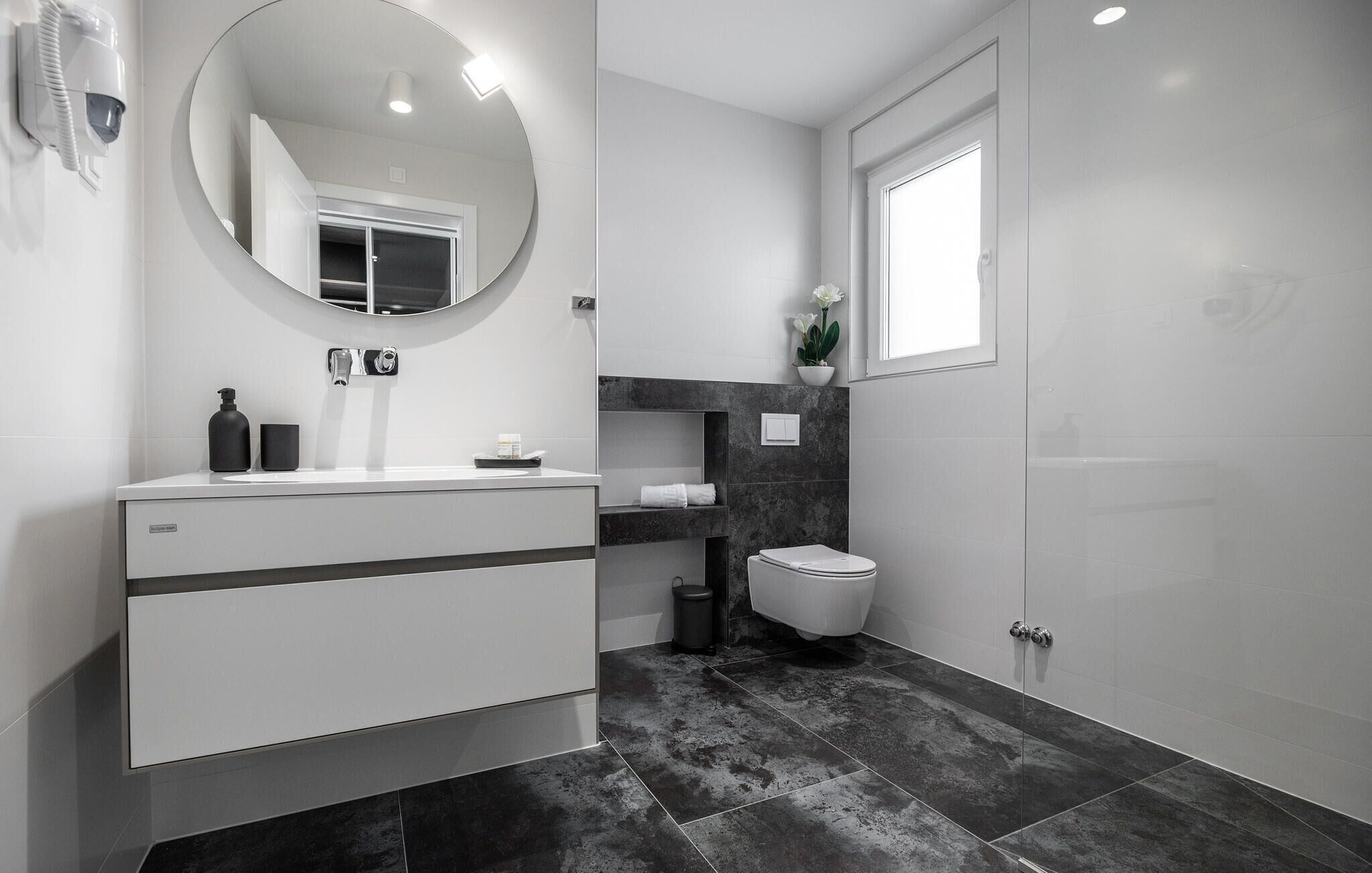 A modern bathroom with a white vanity, a round mirror, a toilet, and a shower area.