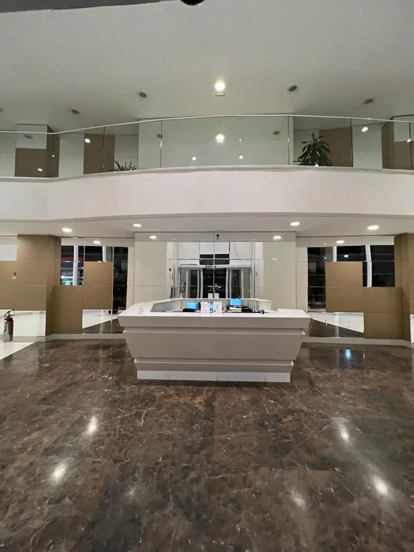 Um hall amplo com um balcão de recepção central, guarda-corpo de vidro no nível superior e um extintor de incêndio no chão.