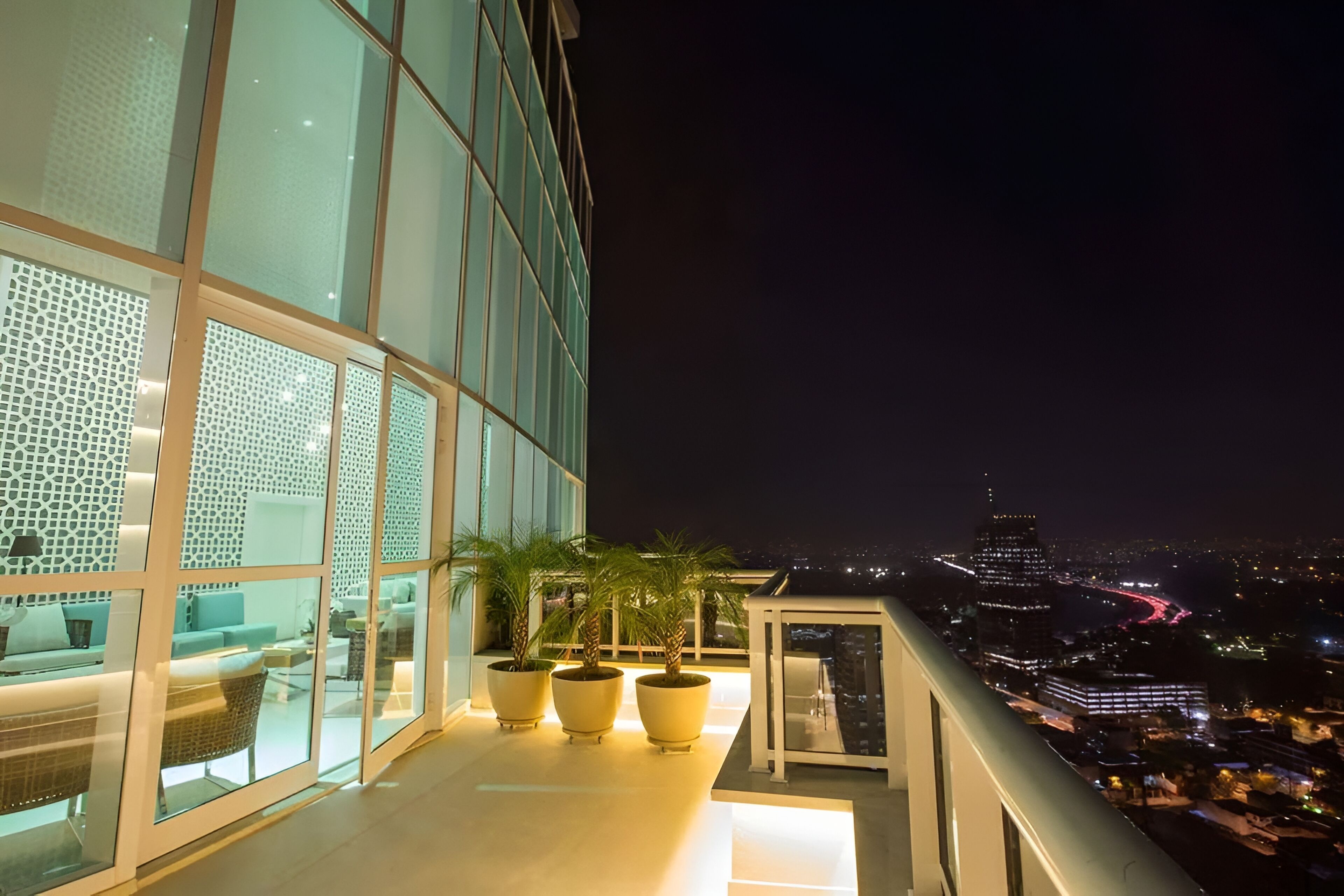 Un bâtiment moderne doté de grandes fenêtres en verre, d’une terrasse avec des sièges et de plantes en pot sur un balcon.