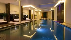 A modern indoor swimming pool with a long, narrow design, surrounded by seating areas and a well-lit ceiling.