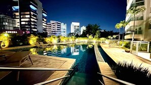 A nighttime view of a swimming pool area with a wooden deck, lounge chairs, and a well-lit pool.