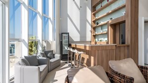 A modern living area with a sofa, a wicker chair, a bar area, and a large window providing natural light.