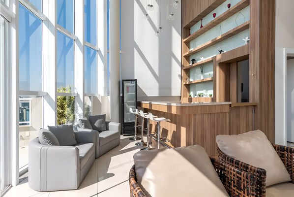 Un espacio de sala moderno con sofá, una silla de mimbre, zona de bar y un amplio ventanal que proporciona luz natural.