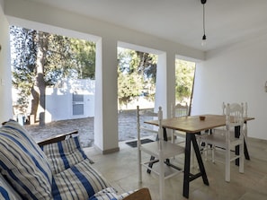 A covered patio area with a wooden table and white chairs, a striped sofa, and a view of trees and a white building.