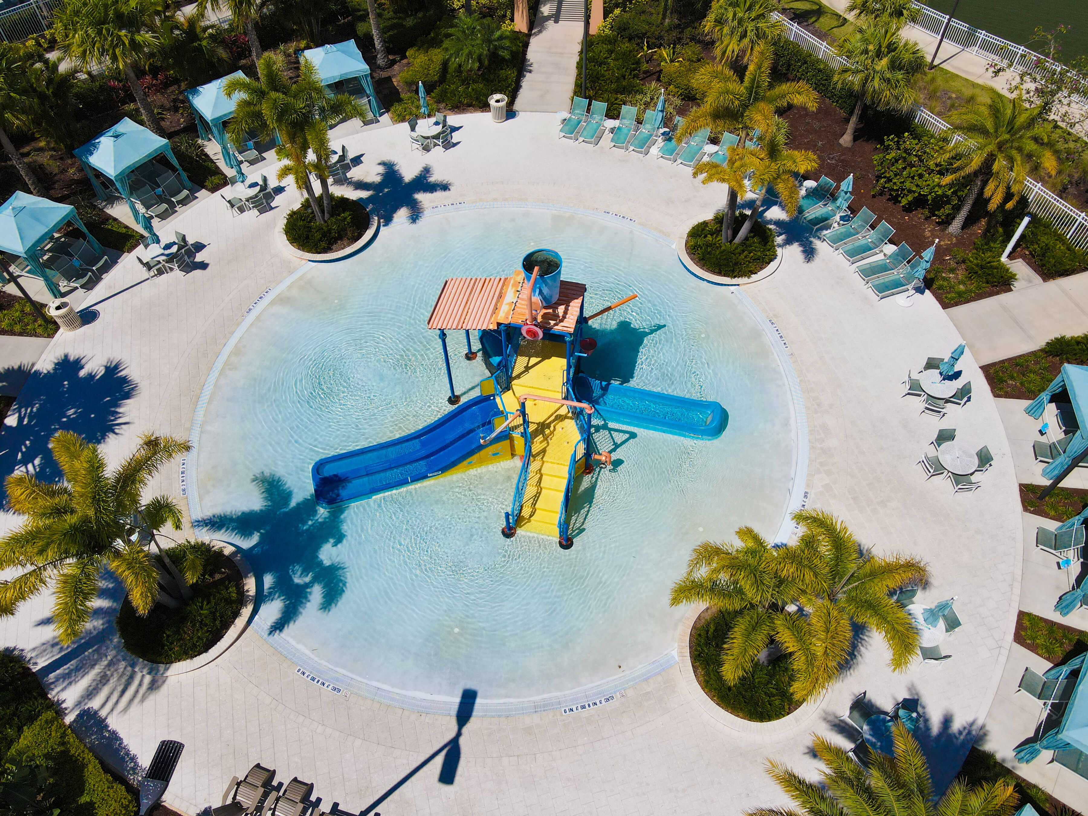 An aerial view of a circular water park with a central slide, surrounded by lounge chairs and palm trees.