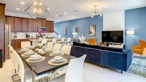 A modern kitchen with wooden cabinets, a dining area with a dark table and white chairs, a blue accent wall, and a TV in the living area.