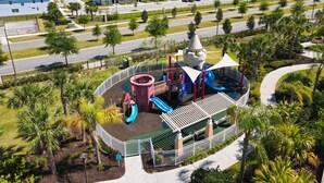 A playground with slides and climbing structures surrounded by a fence, set in a residential area with houses and a road in the background.