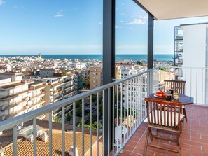 Un balcón con vistas a una ciudad costera, una mesa con frutas y una taza, y una barandilla blanca.