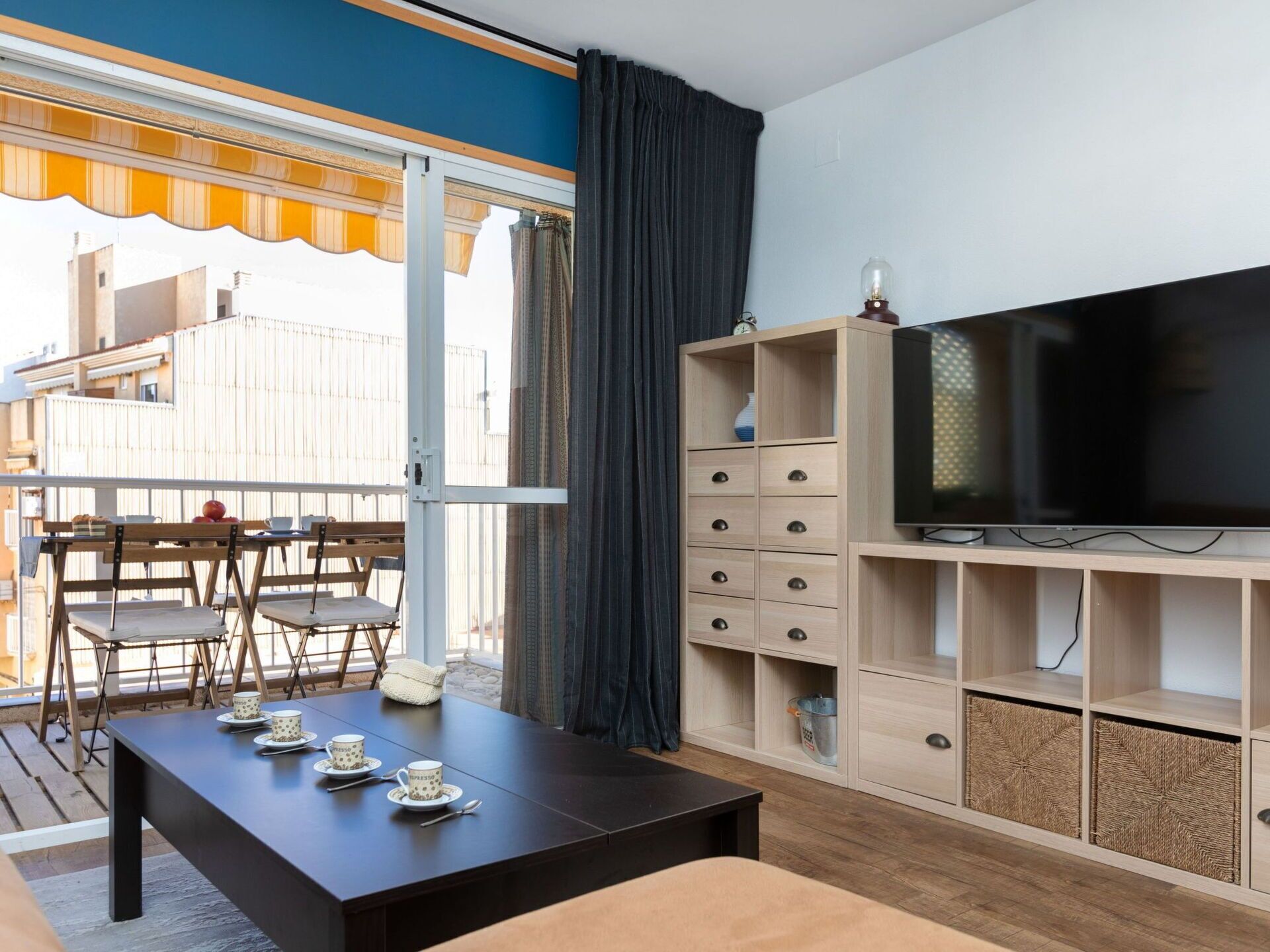 A modern living room with a large flat-screen TV, wooden furniture, and a balcony with a table set for two.