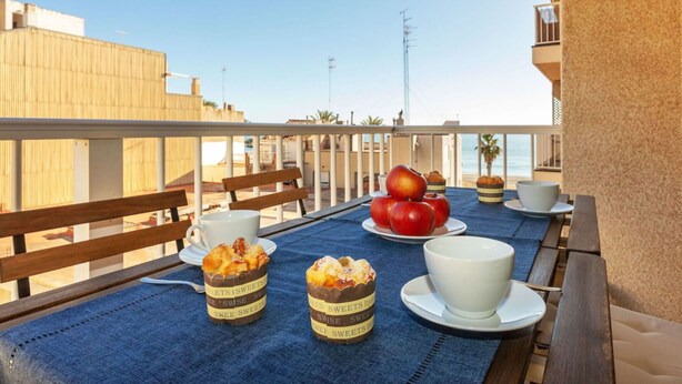 Un tavolo da balcone con vista sul mare, con tovaglia, sedie di legno, una tazza bianca, due piccoli contenitori etichettati 'SWEETS' e un piatto con mele rosse.