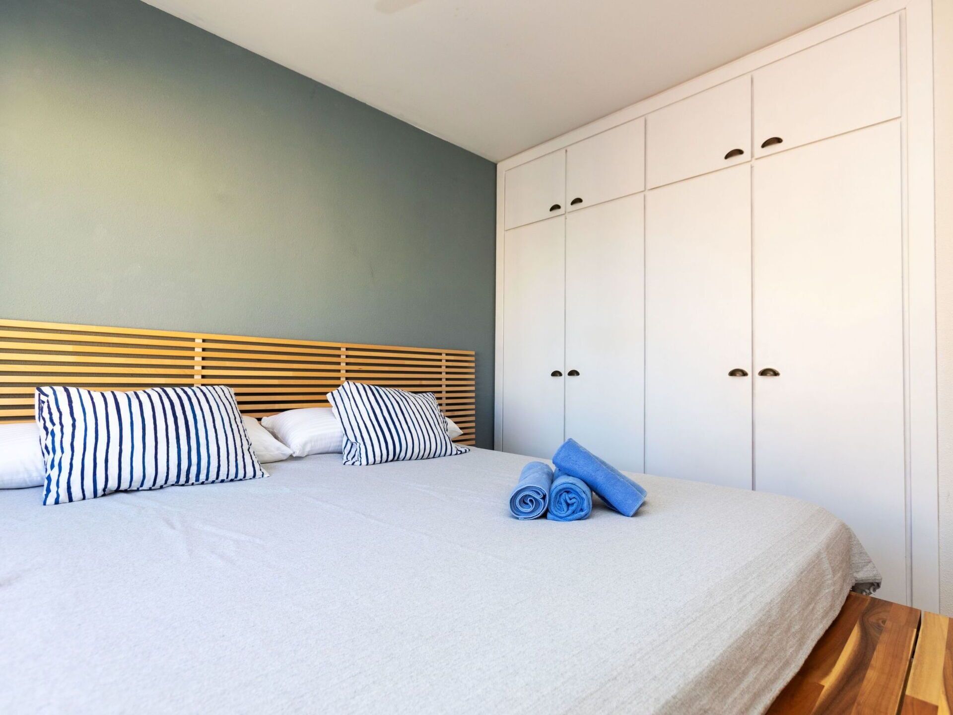 Una cama bien hecha con almohadones a rayas, un cabecero de madera y un armario blanco con varias puertas.