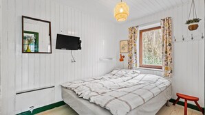 Un dormitorio con cama, televisión, ventana con cortinas, lámpara colgante y un taburete pequeño.