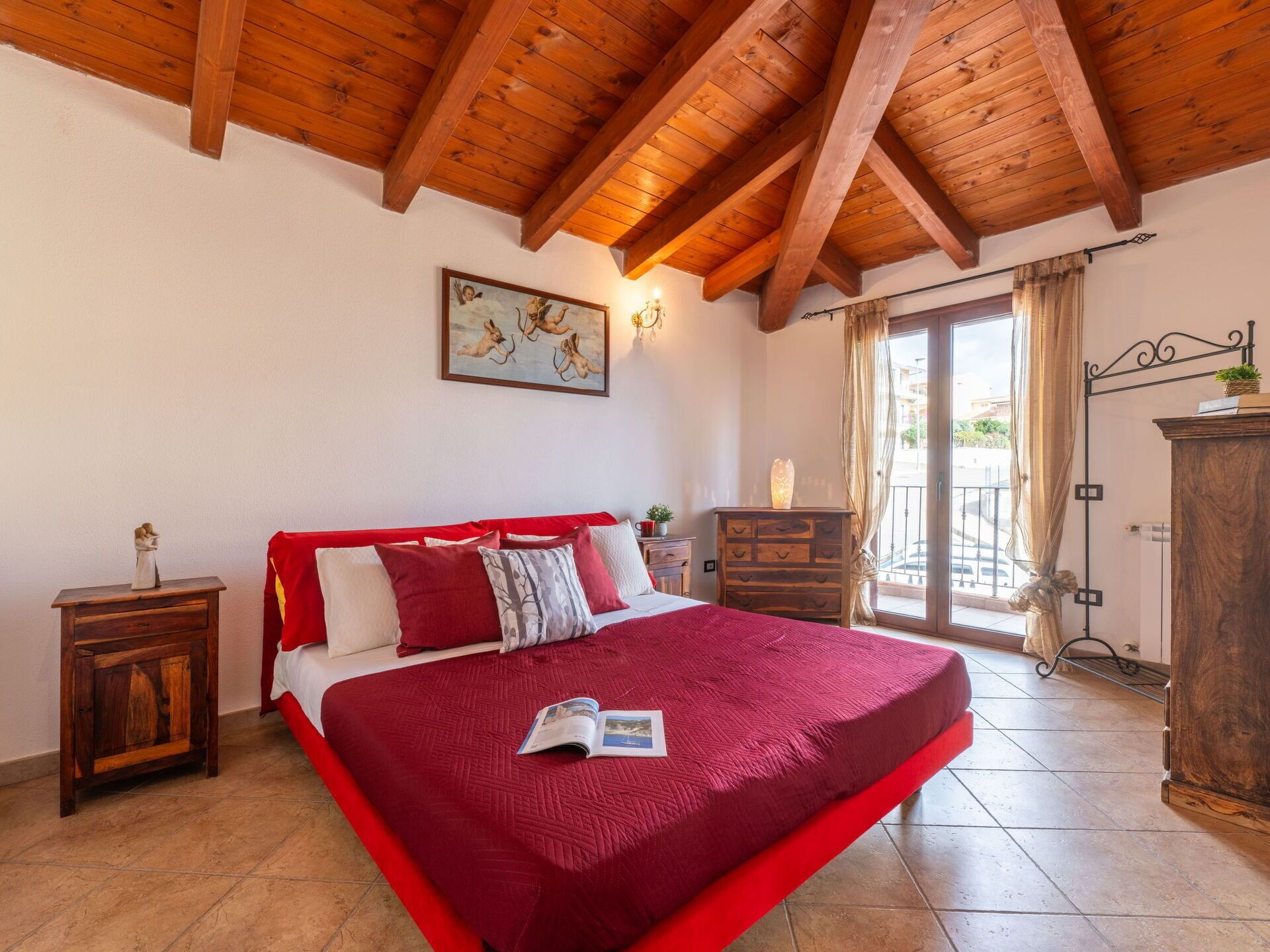 A bedroom with a large bed, wooden ceiling beams, a painting on the wall, and a view through the window.