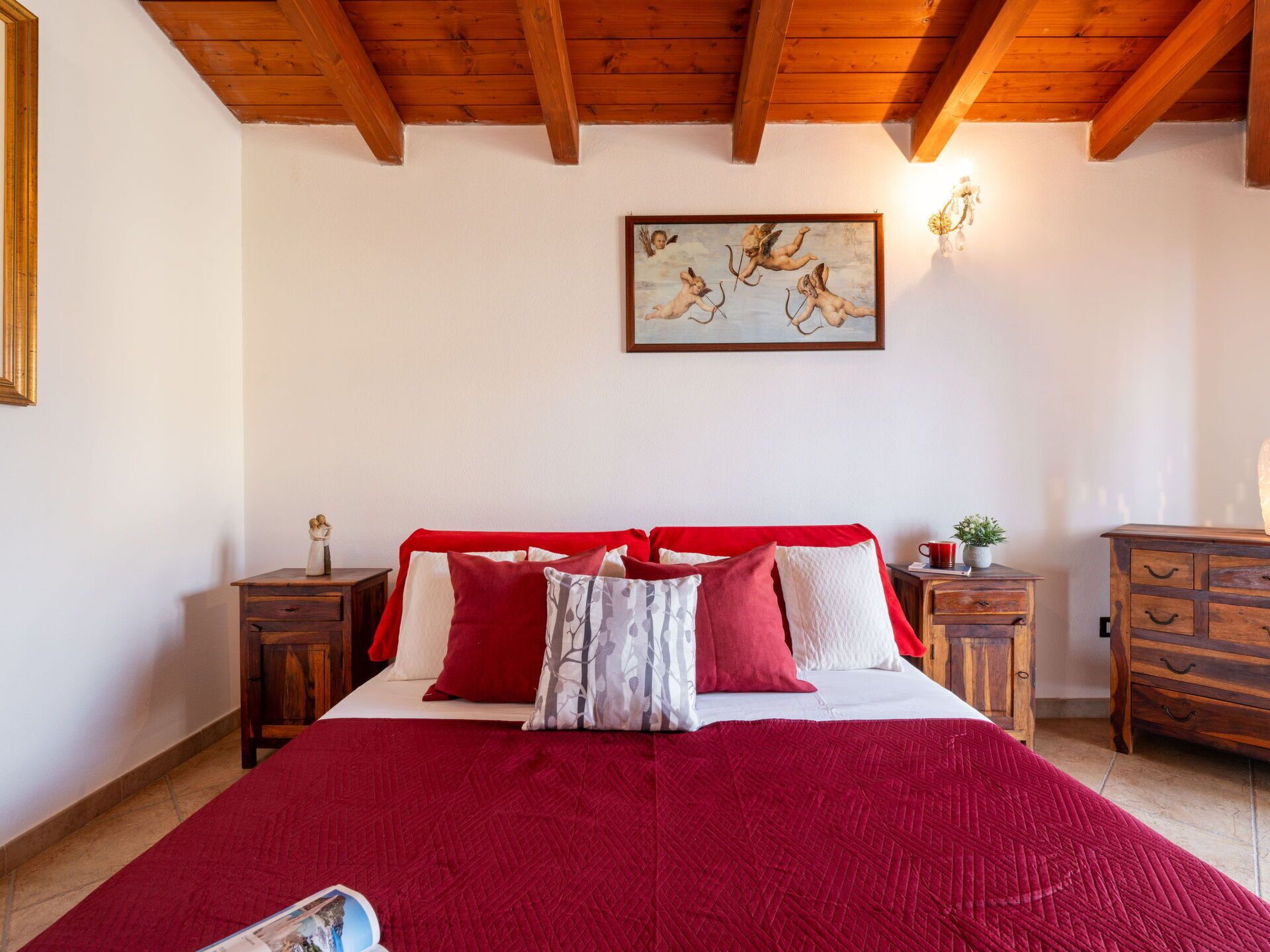 Ein Schlafzimmer mit Holzbalkendecke, einem Bett mit rot-weiß gestreifter Bettwäsche, einem Bild an der Wand und einem Nachttisch mit Lampe.