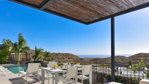 A terrace with a pool, white furniture, and a view of the hills and ocean.