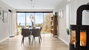 Ein modernes Wohnzimmer mit großem Fenster, einer Essecke mit zwei Stühlen, einem Bücherregal und einem Kaminofen.