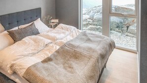 A bedroom with a bed, a nightstand, a vase with flowers, and a view of rocky terrain through large windows. - Stunning home in Stabbestad (Stabbestad)