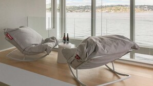 A modern room with two grey rocking chairs, a small table with bottles and glasses, and a large window overlooking a body of water and distant buildings. - Stunning home in Stabbestad (Stabbestad)