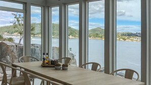 A dining area with a table set for four, large windows with a view of a lake and hills, and a stone wall to the left. - Stunning home in Stabbestad (Stabbestad)