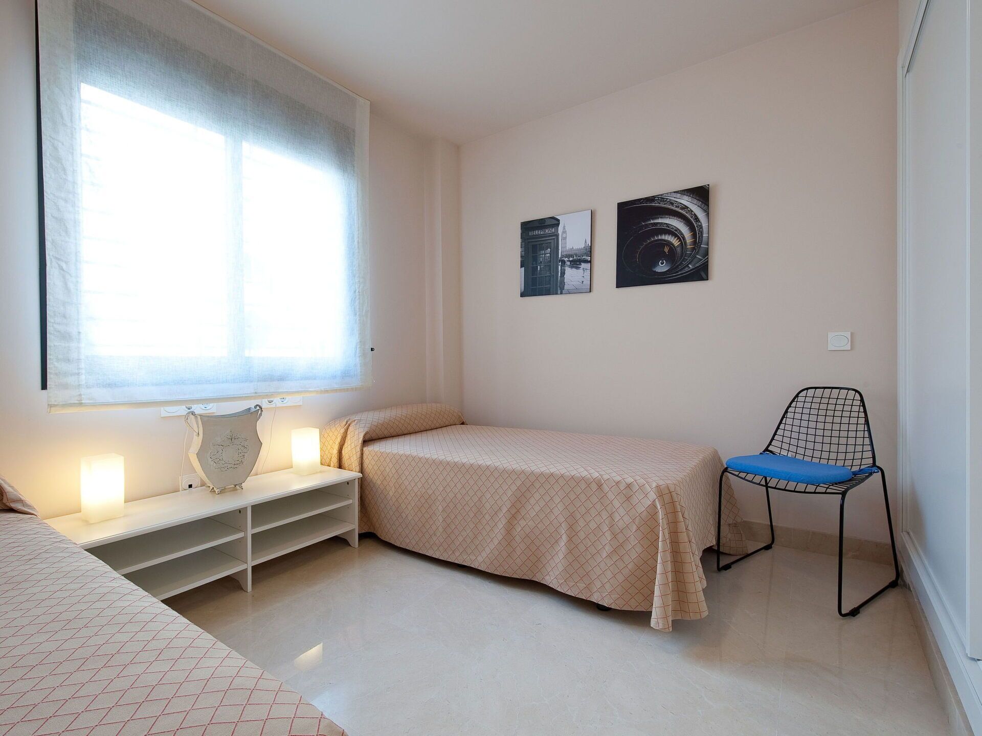 A bedroom with a bed, a chair, a nightstand, a window with blinds, and two wall-mounted pictures.