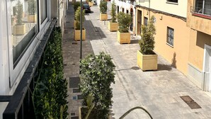 Eine enge Stadtstraße mit Gebäuden an beiden Seiten, Blumentöpfen entlang des Gehwegs und einem klaren, blauen Himmel.