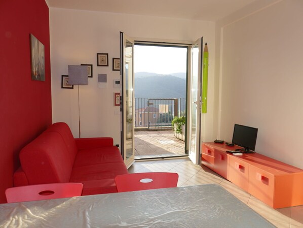 A modern living room with a red sofa, a small table, and a TV on a wooden cabinet. A glass door leads to a balcony with a view of the mountains. - Cosy apartment for 4 people with A/C, WIFI, pool, TV and balcony (Civezza)