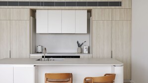 Une cuisine moderne avec un îlot central, deux chaises en bois, une machine à café et une bouilloire posées sur le plan de travail.