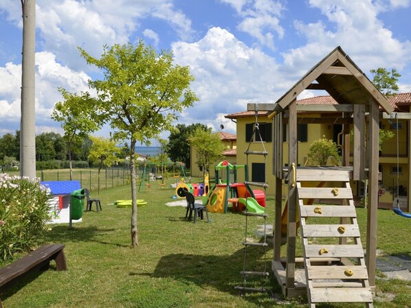 A playground with climbing structures and swings, surrounded by a fenced area with trees and a building in the background.