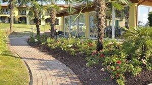 A pathway leading to a building with a yellow facade, palm trees, and a garden with red flowers.