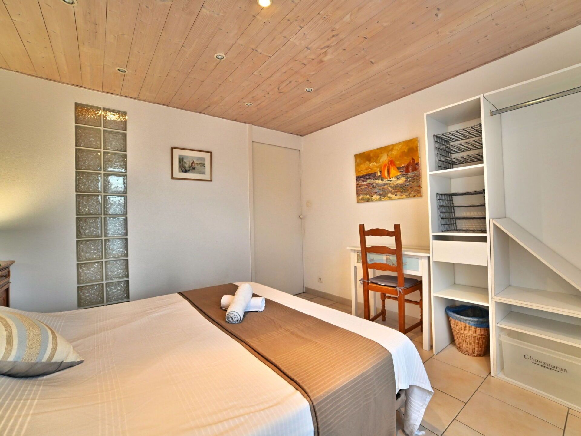 A bedroom with a bed, a desk, and a chair. There is a painting on the wall and a window with a view.