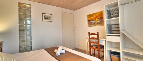 A bedroom with a bed, a desk, and a chair. There is a painting on the wall and a window with a view.