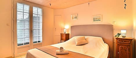 A bedroom with a wooden ceiling, a large bed with a headboard, a bedside table, a nightstand, and a window with blinds.