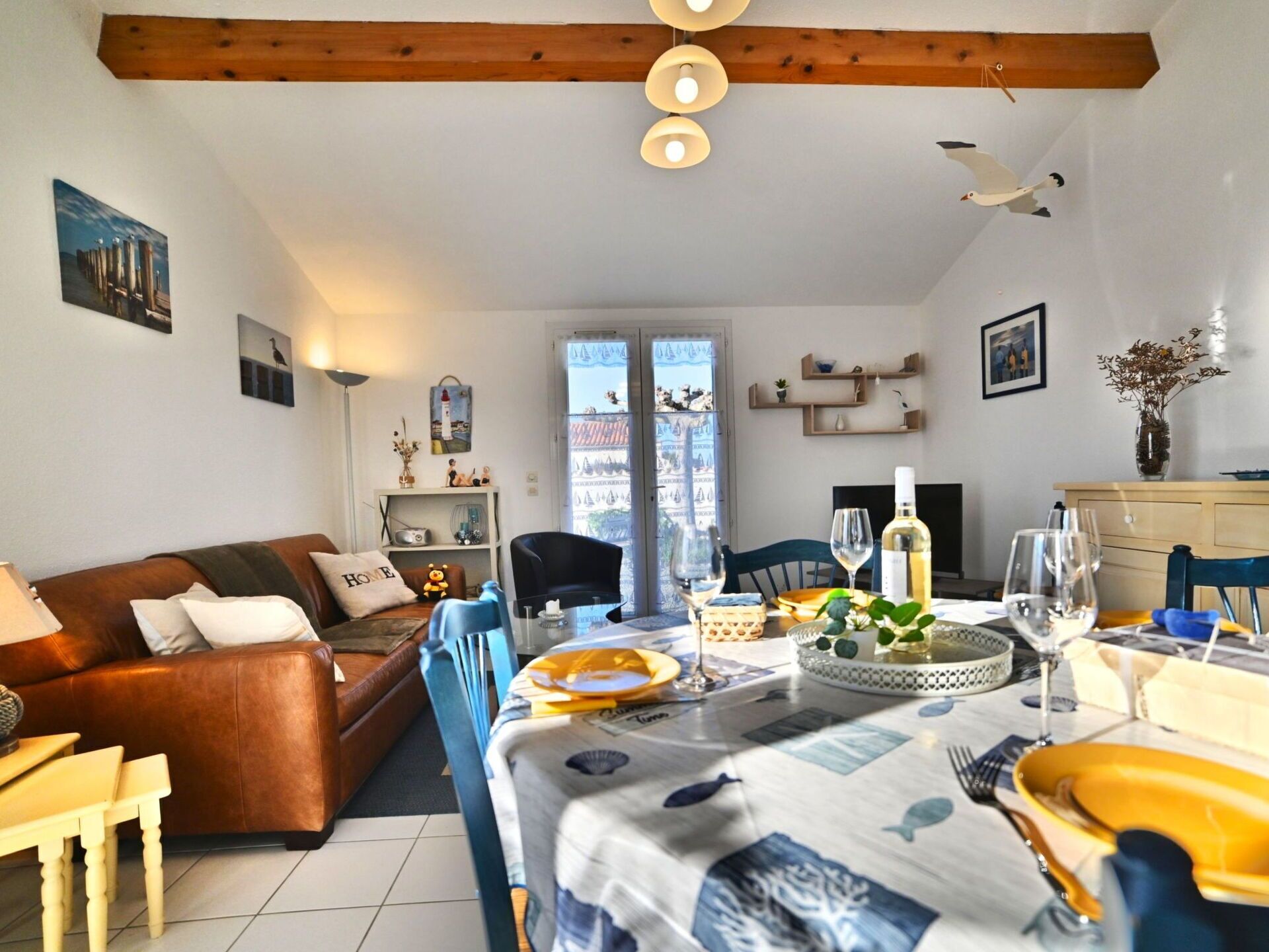 A cozy living room with a brown leather sofa, a dining table set with plates, glasses, and a bottle of wine. There are decorative items on the shelves and a window with a view of buildings.