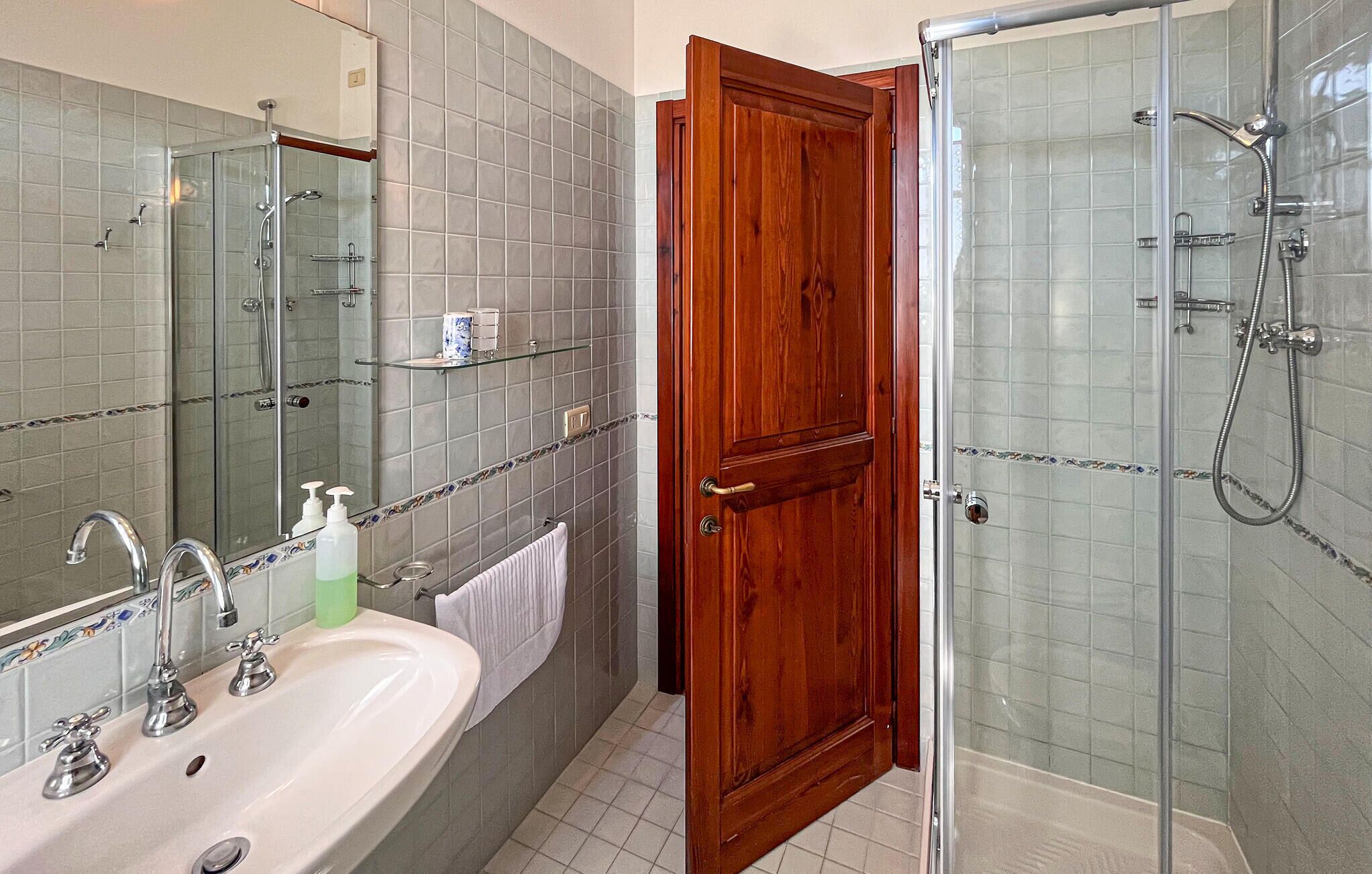 Ein Badezimmer mit Duschkabine aus Glas, einer Holztür und einem weißen Waschbecken mit mehreren Wasserhähnen.