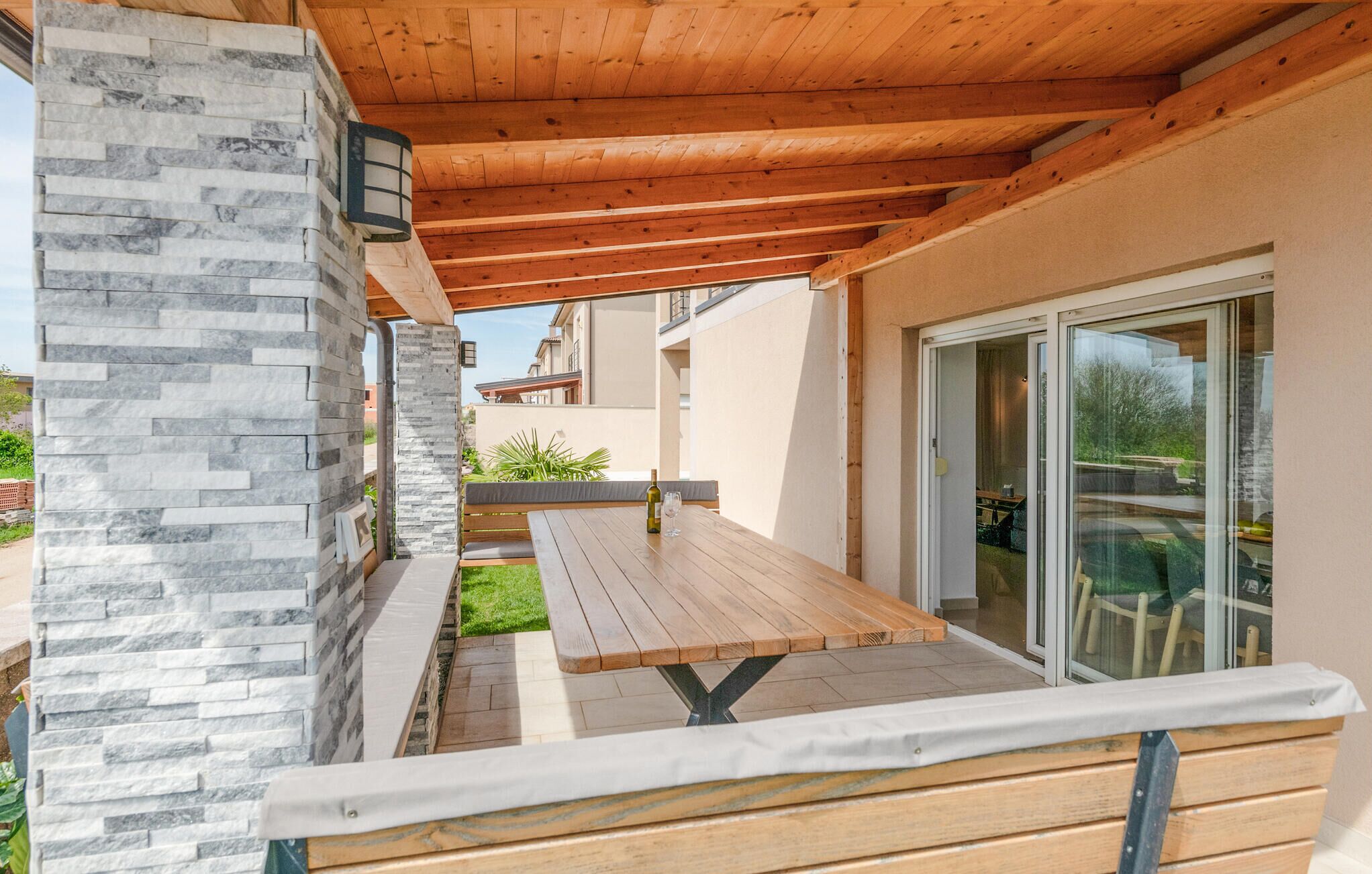 An outdoor patio with a wooden table and benches, a stone wall, and a glass door leading to a garden.