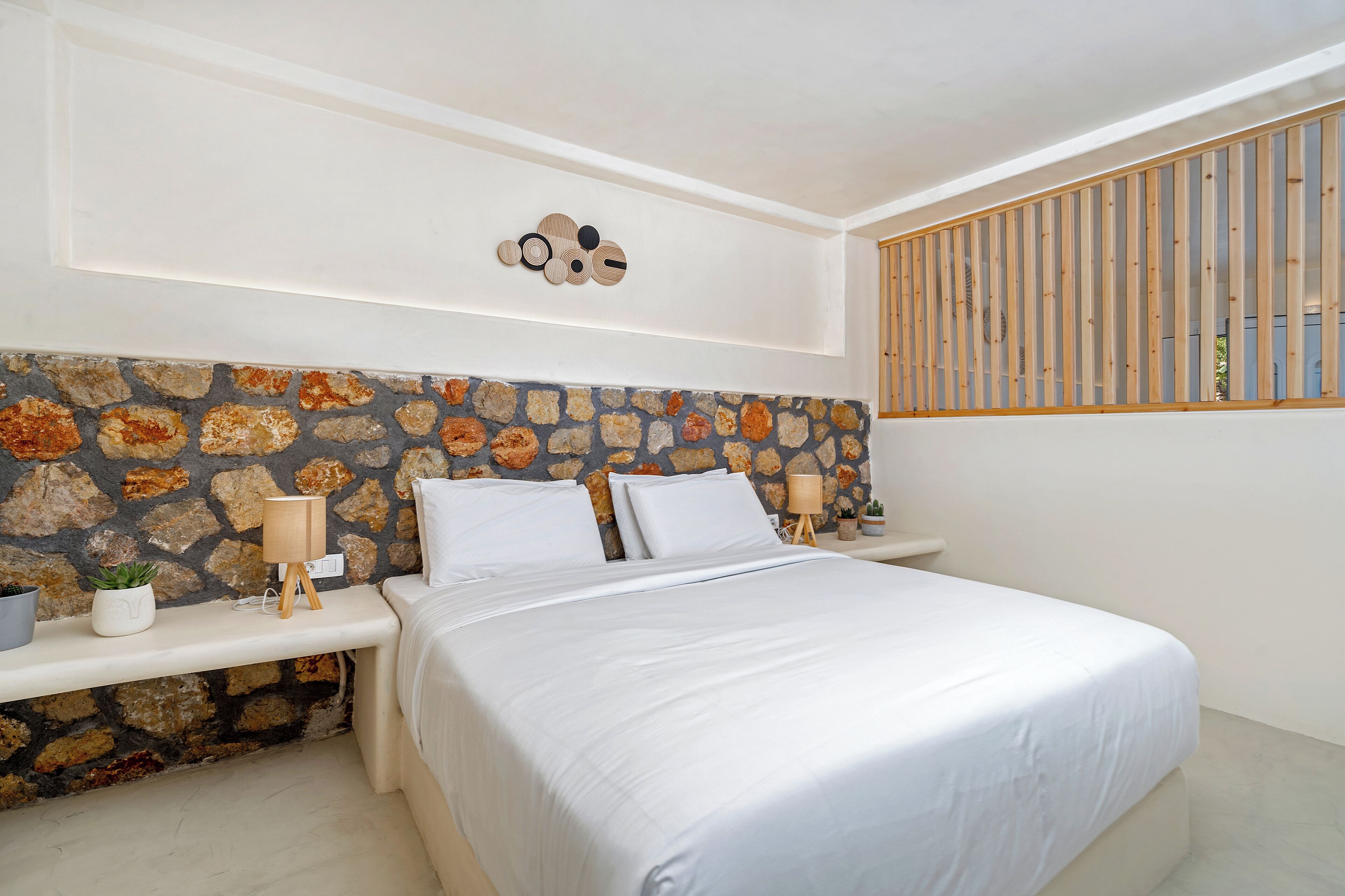 A bedroom with a stone-patterned headboard, a bed with white linens, a bedside table with a lamp, and a window with vertical blinds.