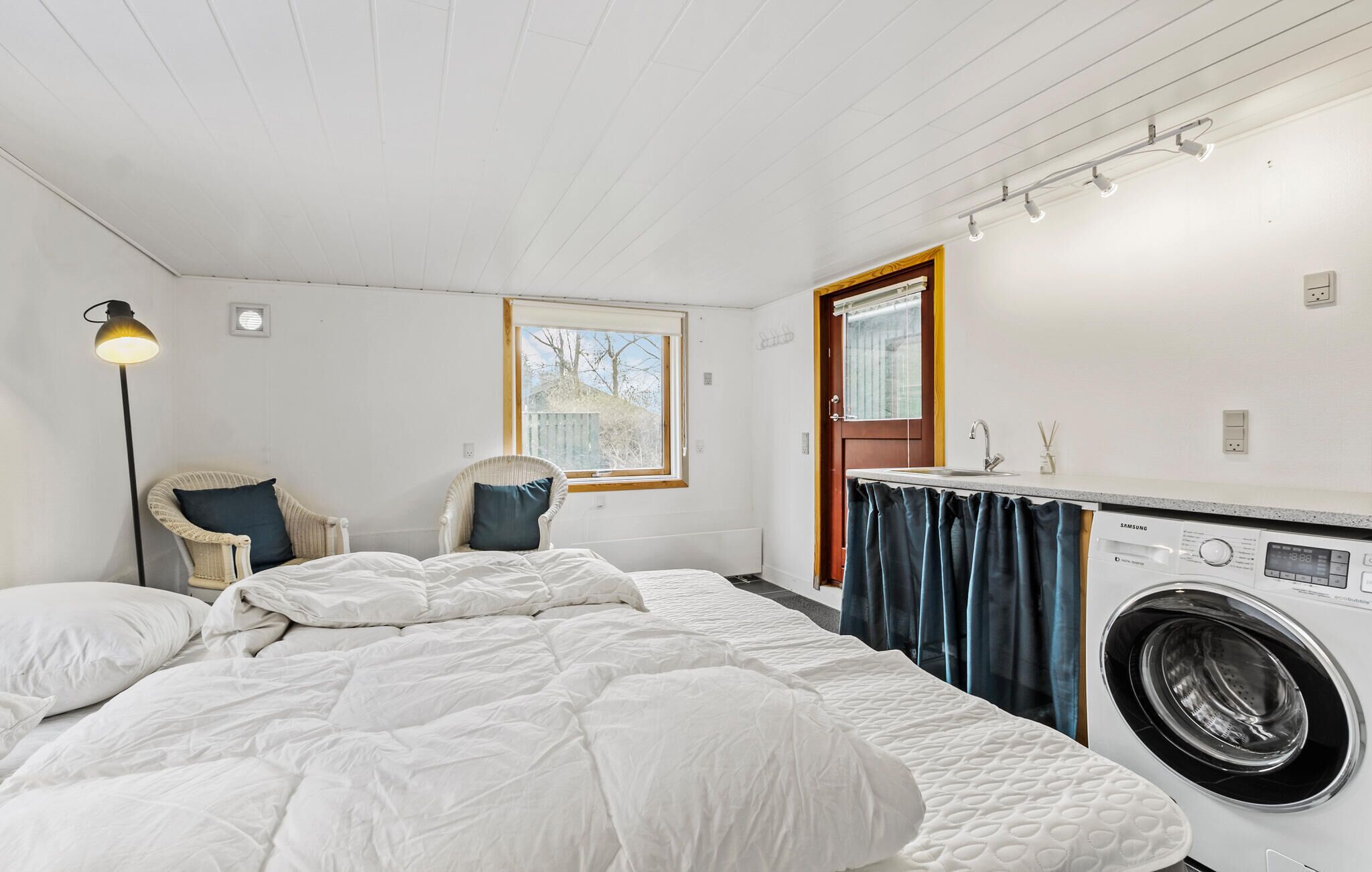 Ein kleines Schlafzimmer mit einem Bett, einer Waschmaschine, einem Sessel und einem Fenster.