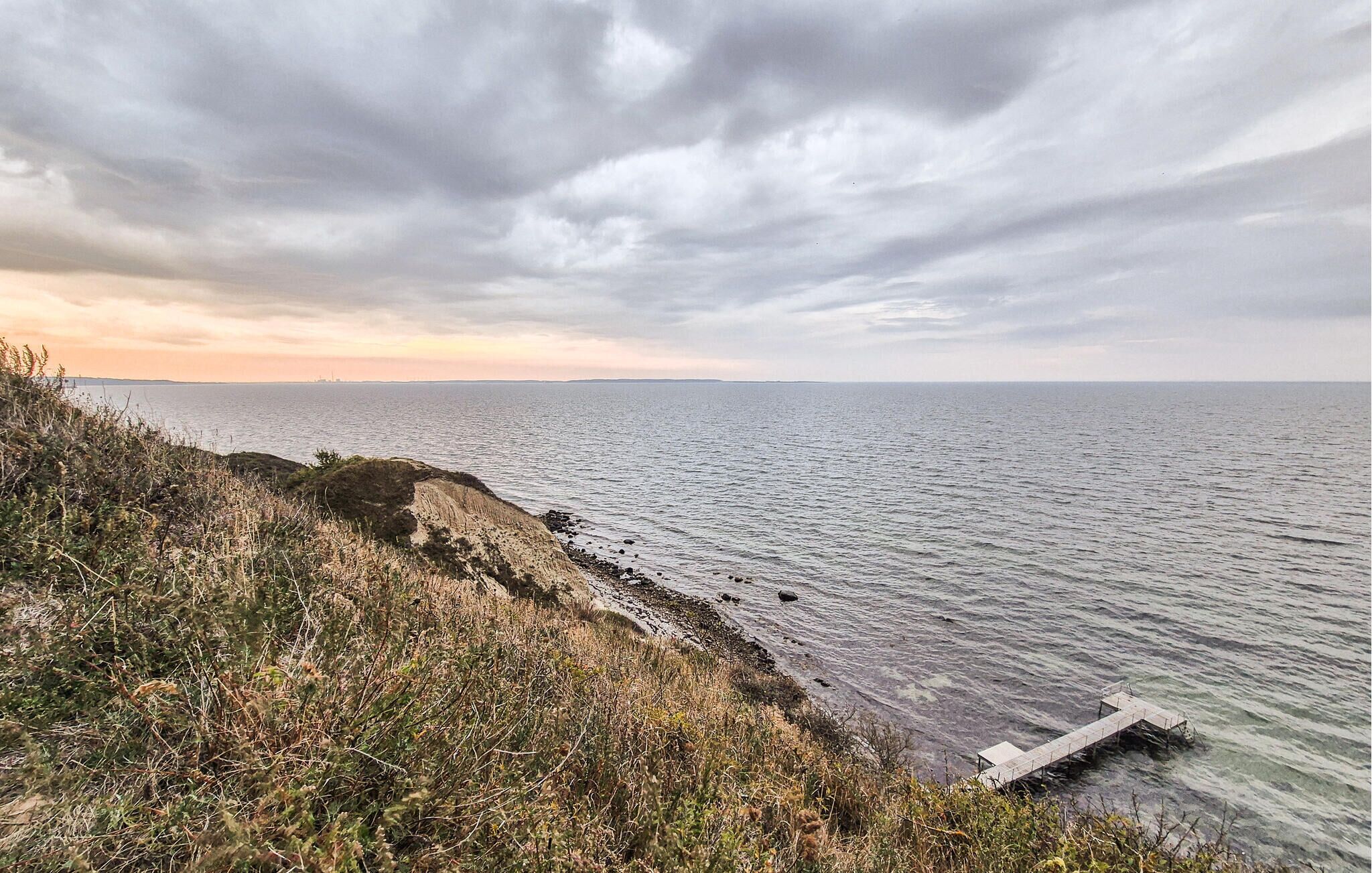 A coastal landscape with a rocky shoreline, a wooden pier extending into the water, and a distant city skyline under a cloudy sky.