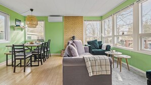 Ein Wohnzimmer mit grüner Wand, einer Wand aus Ziegelsteinen, einem grauen Sofa, einem grünen Sessel, einem Esstisch mit Stühlen und großen Fenstern.