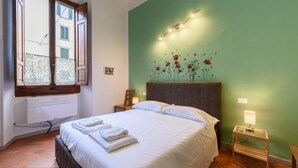Quarto com cama, janela com persianas, parede com um mural floral e mesa de cabeceira com luminária.