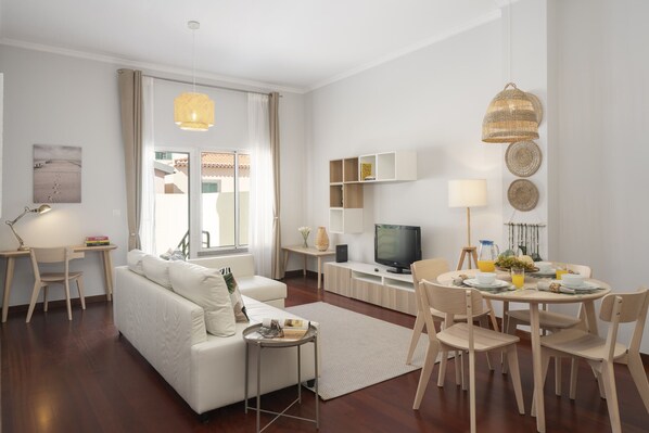A modern living room with a white sofa, dining table, and chairs, a TV, and a large window with curtains.