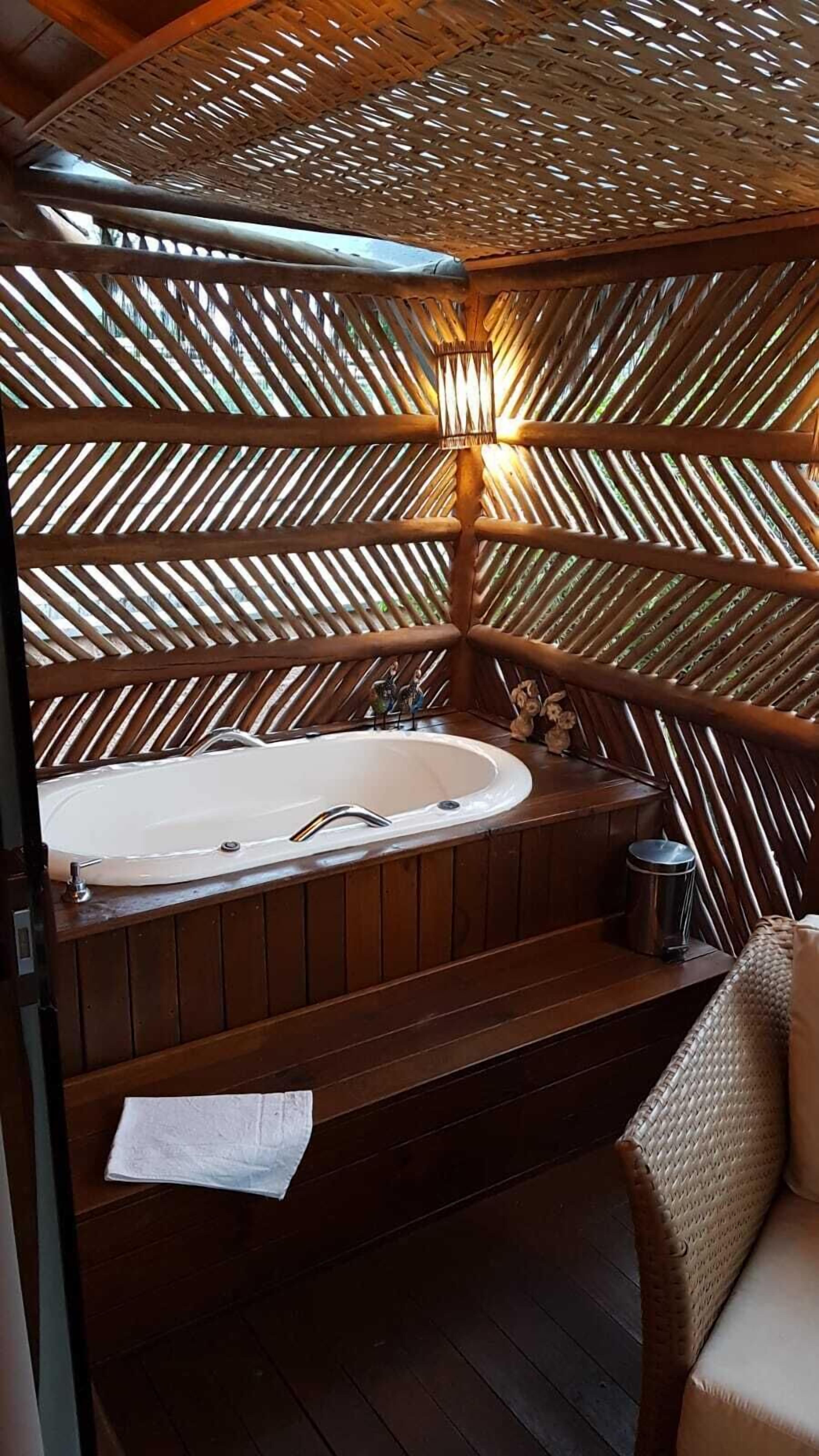 Una habitación con estructura de madera, una bañera grande, una mesita y una silla de mimbre.