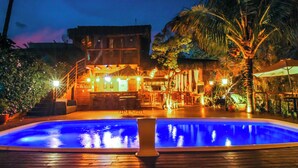 A nighttime view of a pool area with a wooden deck, a bar area with outdoor seating, and a building with multiple windows and a thatched roof. - Pousada de Charme Emirados (Ilhabela)
