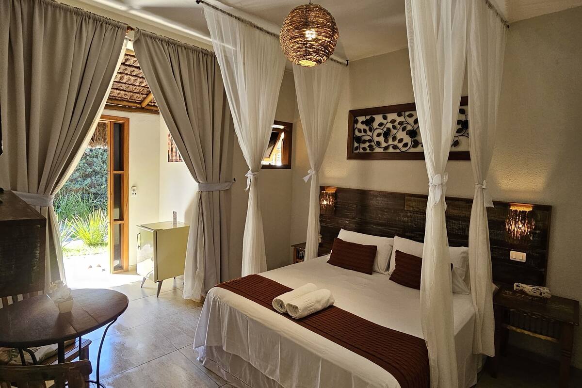 A bedroom with a bed, a bedside table, a chair, a table, and a hanging light fixture.